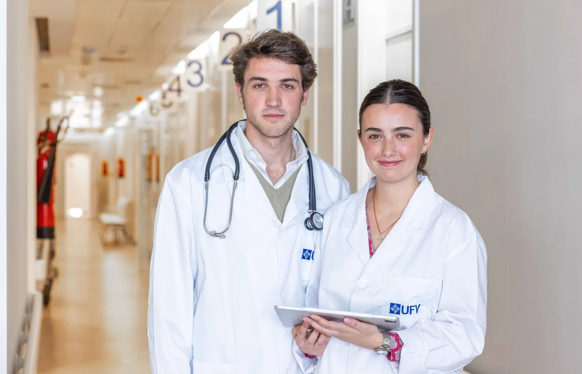 Dos estudiantes de Medicina, con bata y estetoscopio, recorren un pasillo del campus